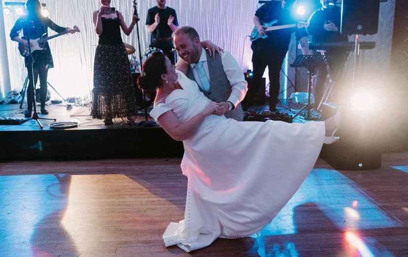 A white couple on their wedding day during their First Dance.  They are doing a 'dip pose'.  The bride is in a white dress with cap sleeves, her brown hair is neatly styled into a low bun and you can just see one white heeled foot under her dress where her leg has come off the floor.  The groom is wearing a light grey waistcoat suit with a white shirt.  They are both smiling at each other.  Behind them you can see their music band