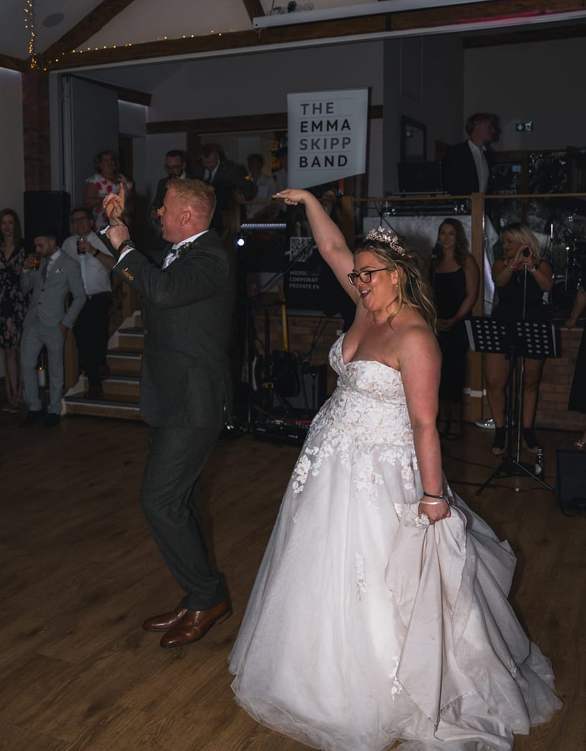 A white couple dancing side by side on their wedding day.  The bride is in a white strapless dress, holding the train with her left hand, whilst her right arm is in the air.  She has glasses and a tiara in her hair.  The groom is in a dark suite with brown shoes, he is in mid dance pose.  Behind them is a banner that reads 'The Emma Skip Band' and in the background are their wedding guests.   They are both smiling 