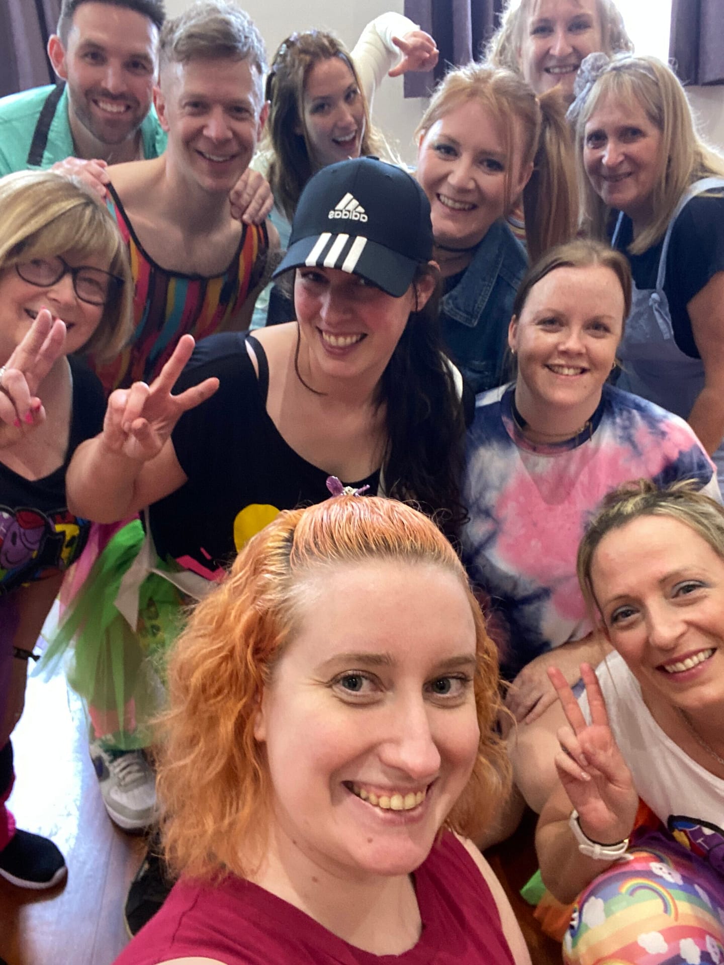 A group of people huddled together, at the end of a dance class, wearing brightly coloured 90's themed gym gear.  They're all smiling at the camera.  Charlotte, a white person with ginger curly hair is at the foreground of the picture